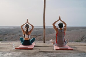 Deux personnes pratiquent le yoga sur des tapis à l’extérieur, avec une vue panoramique sur des collines en arrière-plan.