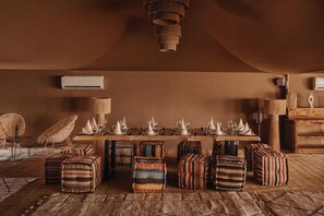 A dining area with a long table set for a meal, surrounded by upholstered ottomans and a woven rug. - Mia Agafay Resort (Agafay)