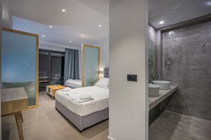 Una habitación de hotel moderna con cama, mesitas de noche, un escritorio y un baño con un espejo grande y dos lavamanos.