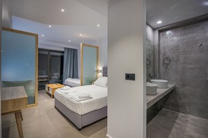 Quarto de hotel moderno com cama, mesas de cabeceira, escrivaninha e banheiro com espelho grande e duas pias.