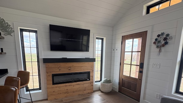 A modern living room with a fireplace, a flat-screen TV, wooden flooring, and large windows.