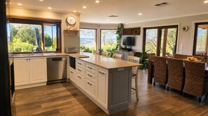 A modern kitchen with white cabinetry, a central island, and a dining area with wicker chairs. - Brindabella Country Escape | Relax, Unwind & Explore (Uriarra)