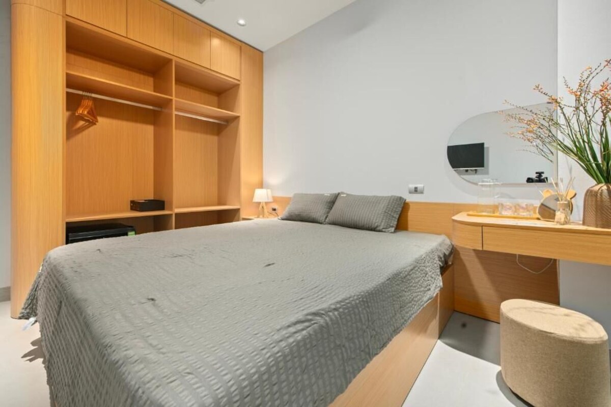 A bedroom with a bed, a wooden wardrobe, a vanity with a mirror, and a small round ottoman.