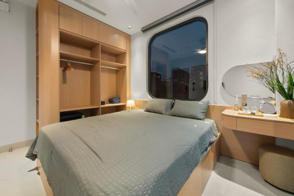 A modern bedroom with a large bed, built-in wooden wardrobe, a bedside table with a lamp, and a view of the city through a window.