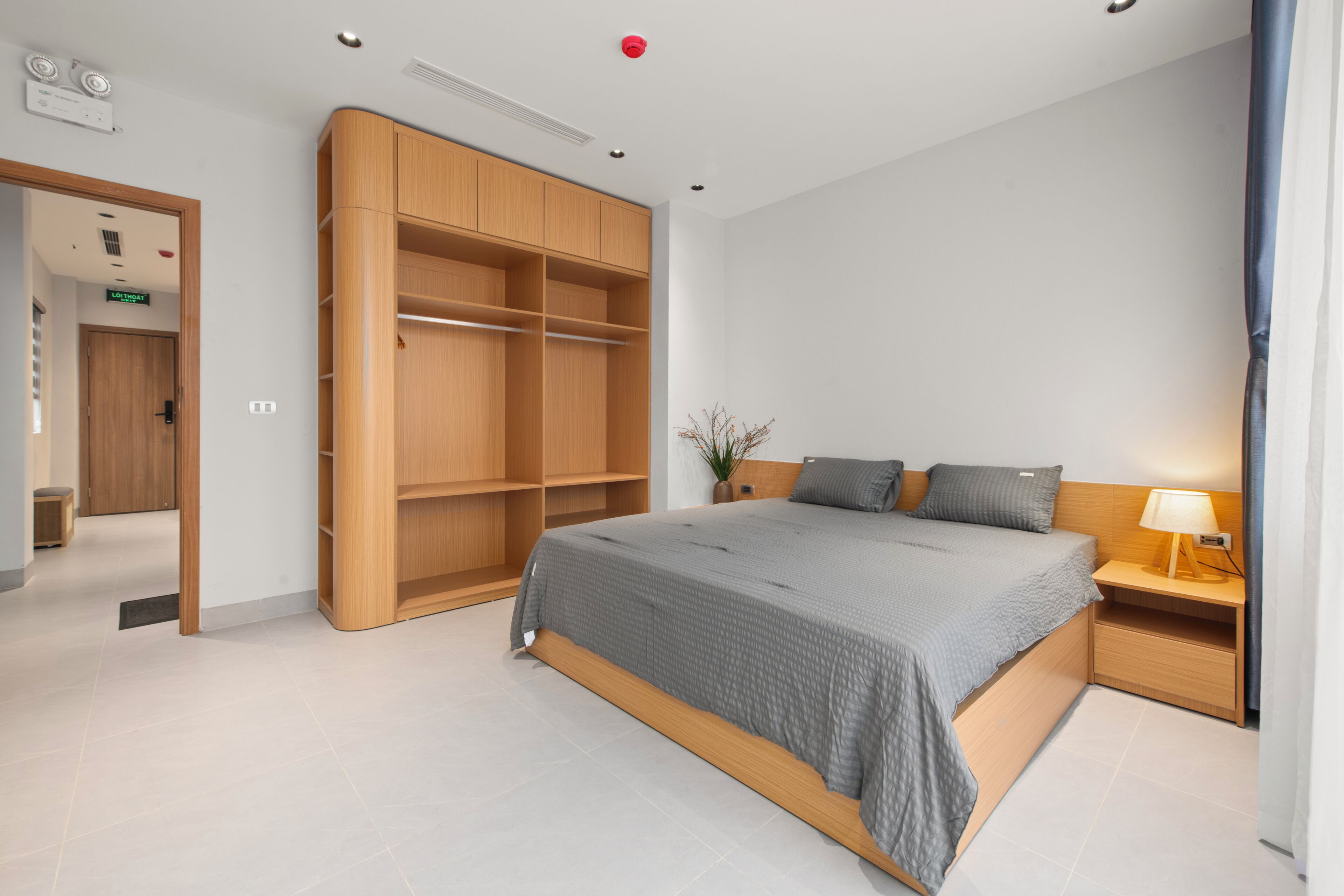 A modern hotel room with a large bed, built-in wooden wardrobe, bedside table with lamp, and a small potted plant.