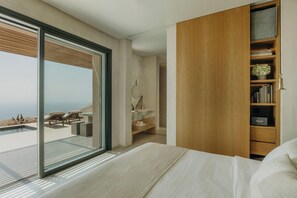 A modern bedroom with a large bed, a wooden wardrobe, a bathroom with a marble countertop, and a balcony with outdoor furniture. - Santorini Sky (Santorini)