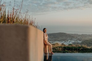 A person sitting on a ledge overlooking a body of water with distant hills. - Santorini Sky (Santorini)