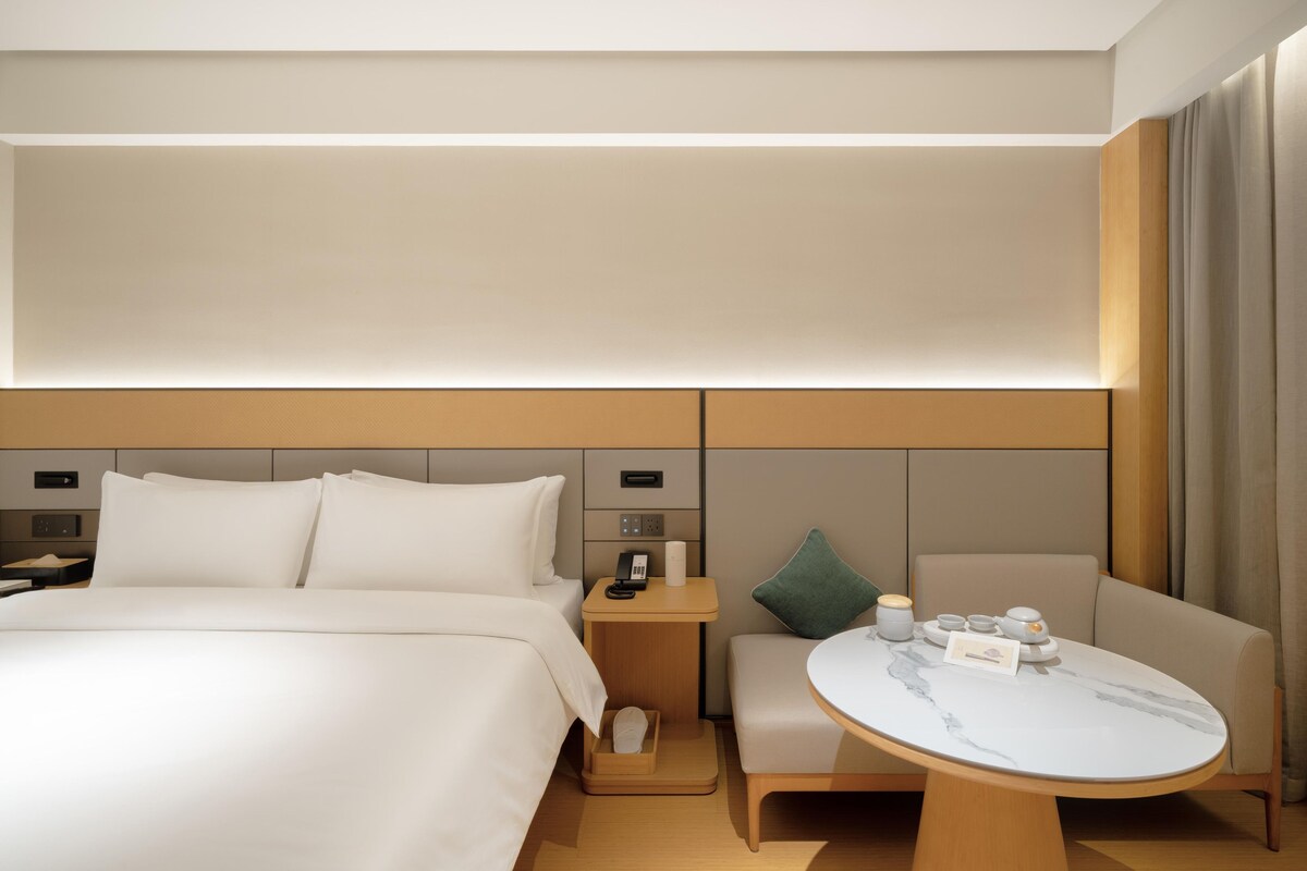 Ein modernes Hotelzimmer mit einem großen Bett, einem Nachttisch, einem kleinen Schreibtisch, einem Sessel und einem runden Tisch mit Marmorplatte.