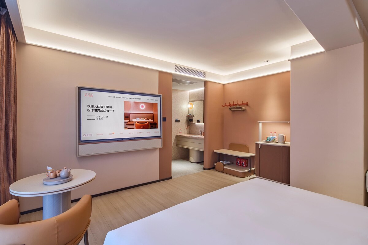 A hotel room with a bed, a small table with a teapot, a chair, a TV mounted on the wall, and a bathroom visible through an open door.