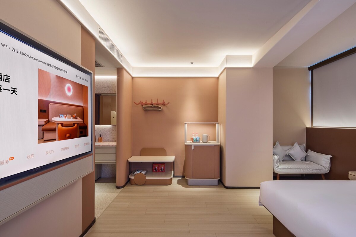 A hotel room with a bed, a desk, a chair, a small table with a mirror, and a TV screen displaying a hotel advertisement.
