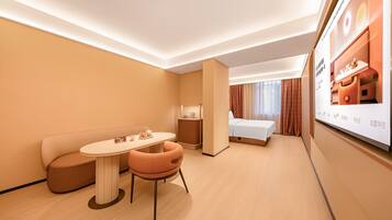 Quarto de hotel com uma cama, área de estar com uma mesa, televisão e mesa de cabeceira com abajur.