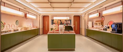 Un café moderno y bien iluminado con una barra central, armarios verdes y diversos productos alimenticios y bebidas a la vista.