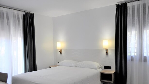 A hotel room with a bed, two bedside tables, a nightstand, and curtains. - Hostal Restaurante Villa De Brihuega (Brihuega)