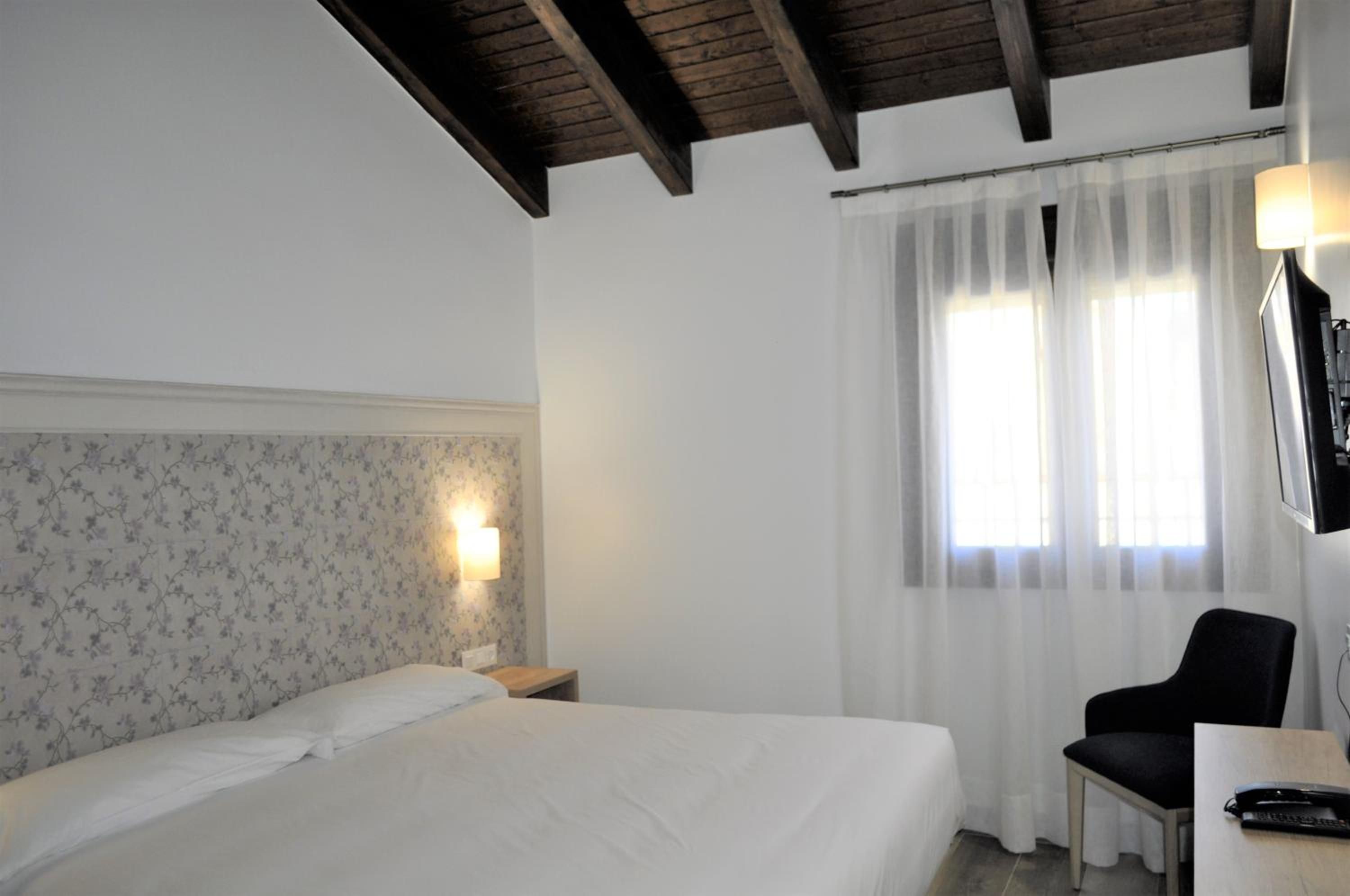 Habitación de hotel con cama, una silla, un escritorio, una lámpara y ventana con cortinas.