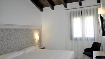 Habitación de hotel con cama, una silla, un escritorio, una lámpara y ventana con cortinas.