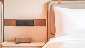 A hotel room with a wooden headboard, a bedside table with a lamp, and a control panel with a speaker.