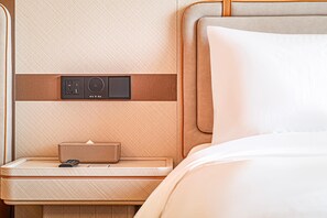 A hotel room with a wooden headboard, a bedside table with a lamp, and a control panel with a speaker.