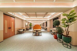 A modern hotel corridor with a central seating area, potted plants, and a clear exit sign.