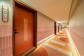 A hotel corridor with a patterned carpet, a wooden door, and a wall-mounted elevator control panel. - Orange Hotel (Xi'an)