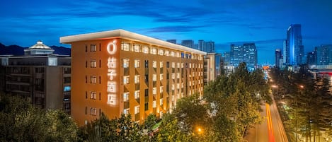 A hotel with a modern design, featuring a prominent 'O' logo on the facade, surrounded by lush greenery and a city skyline in the background.