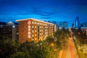 A hotel with a modern design, featuring a prominent 'O' logo on the facade, surrounded by lush greenery and a city skyline in the background. - Orange Hotel (Xi'an)