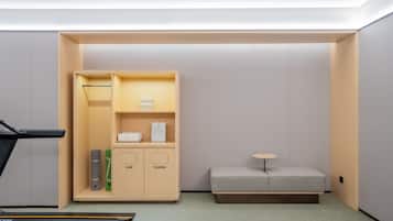 A compact room with a treadmill, a small seating area, and a wardrobe with labeled compartments.