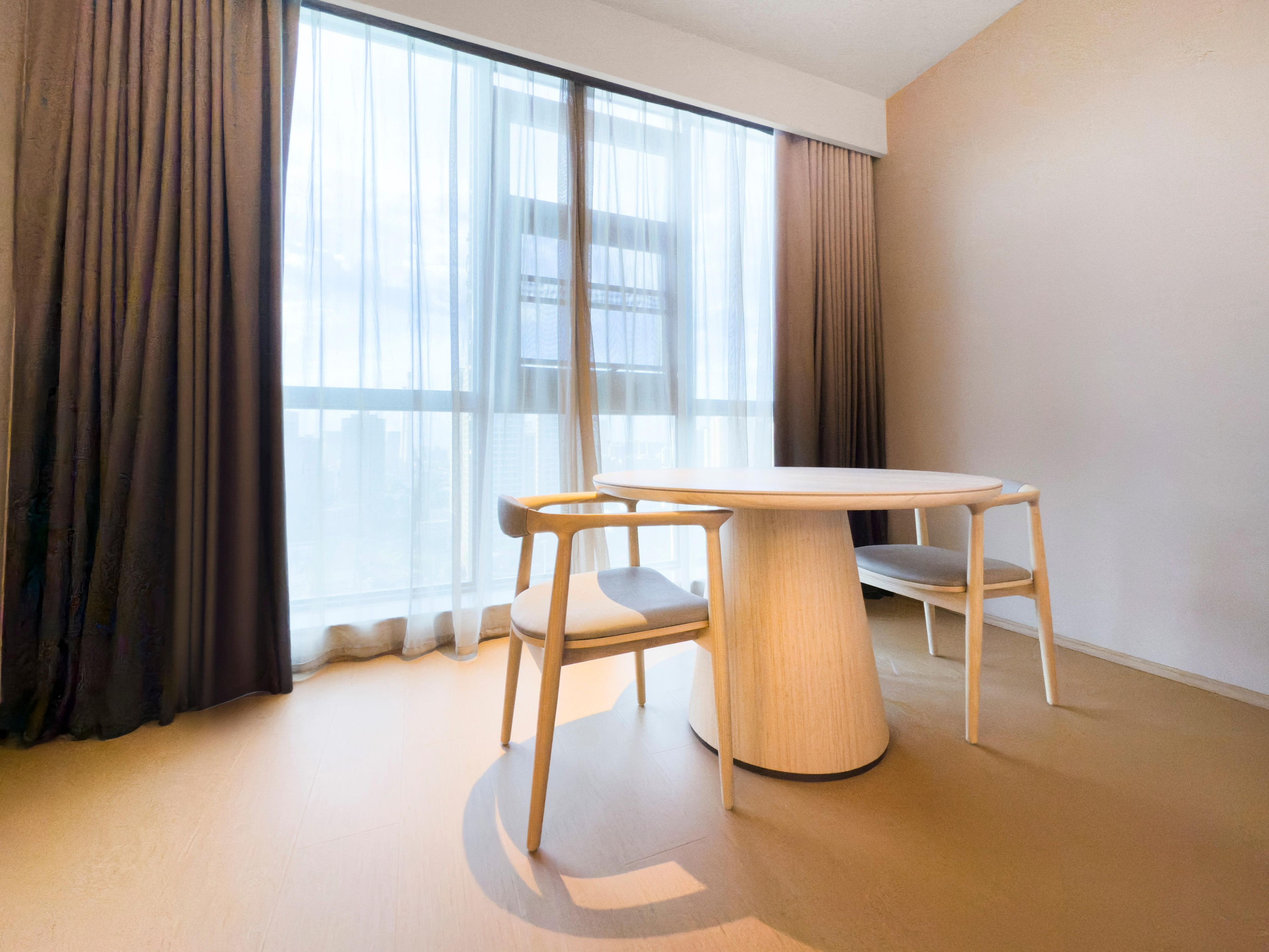 Una habitación moderna con una mesa redonda y dos sillas, ventanas amplias con cortinas y una pared lisa.