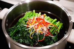 A stainless steel pot containing a dark liquid and green leafy vegetables with thinly sliced red and yellow vegetables on top. - JI Hotel (Xi'an)