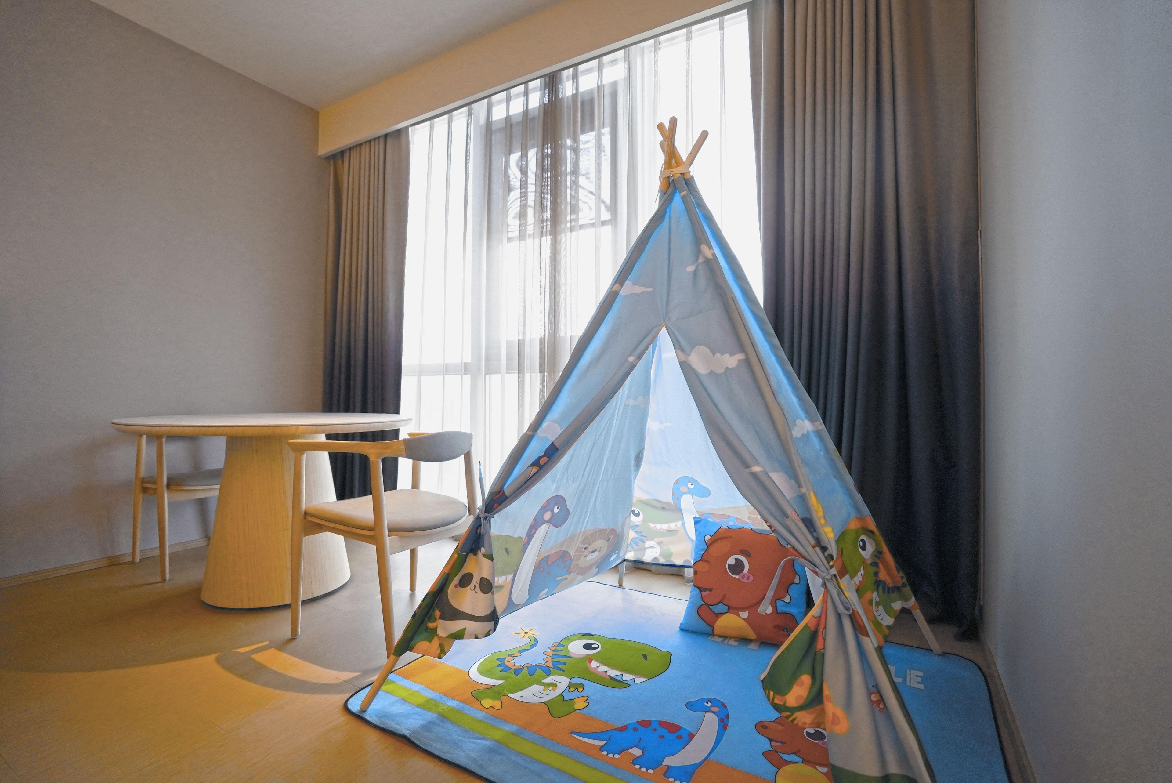 A child's play tent with dinosaur prints, a round table, and a chair in a room with large windows and curtains.