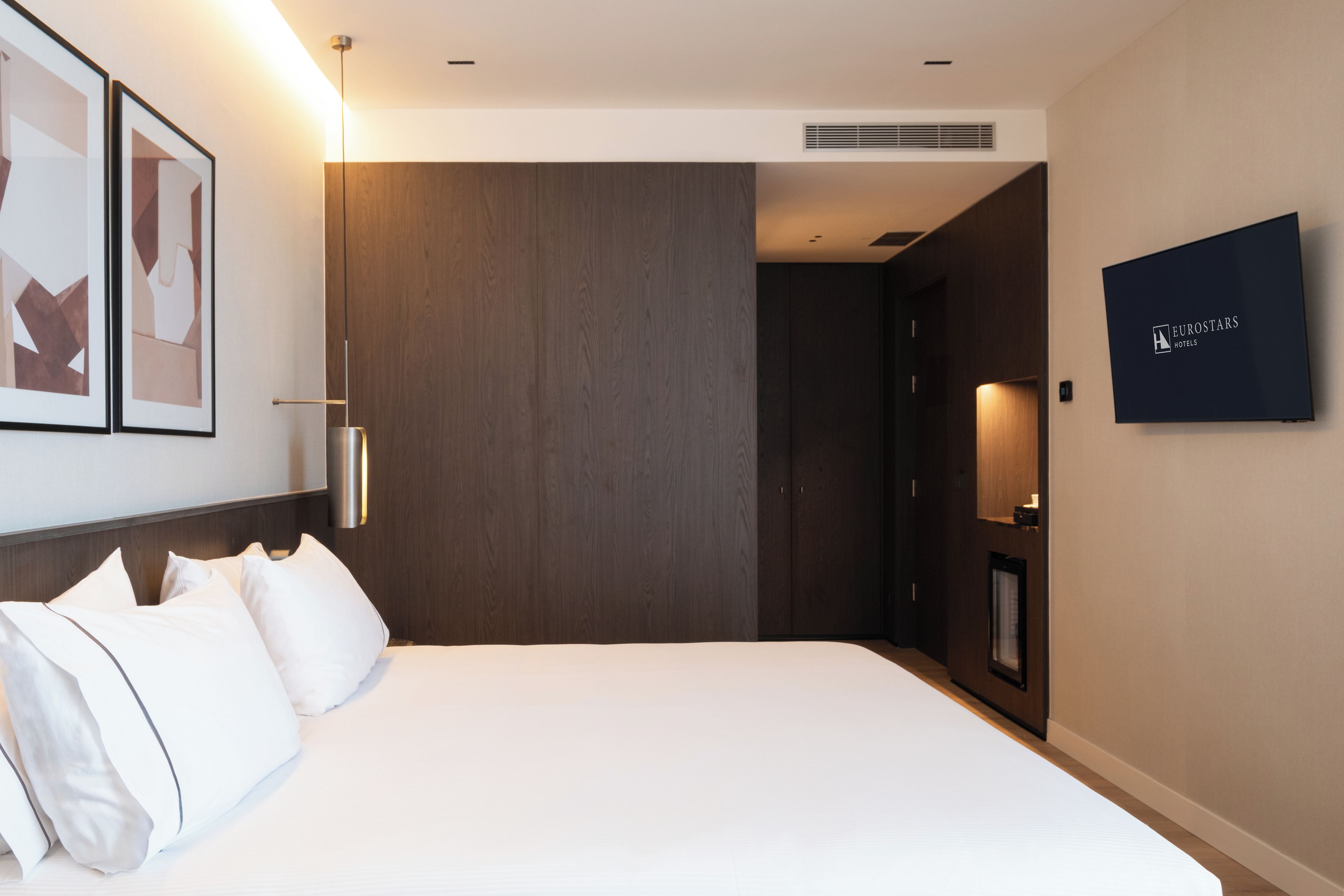 A hotel room with a bed, a wall-mounted TV, and a door leading to another room.