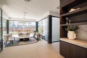 Habitación de hotel moderna con un amplio ventanal, una zona de estar con sofás y una mesa de centro, y un estante de madera con objetos decorativos.