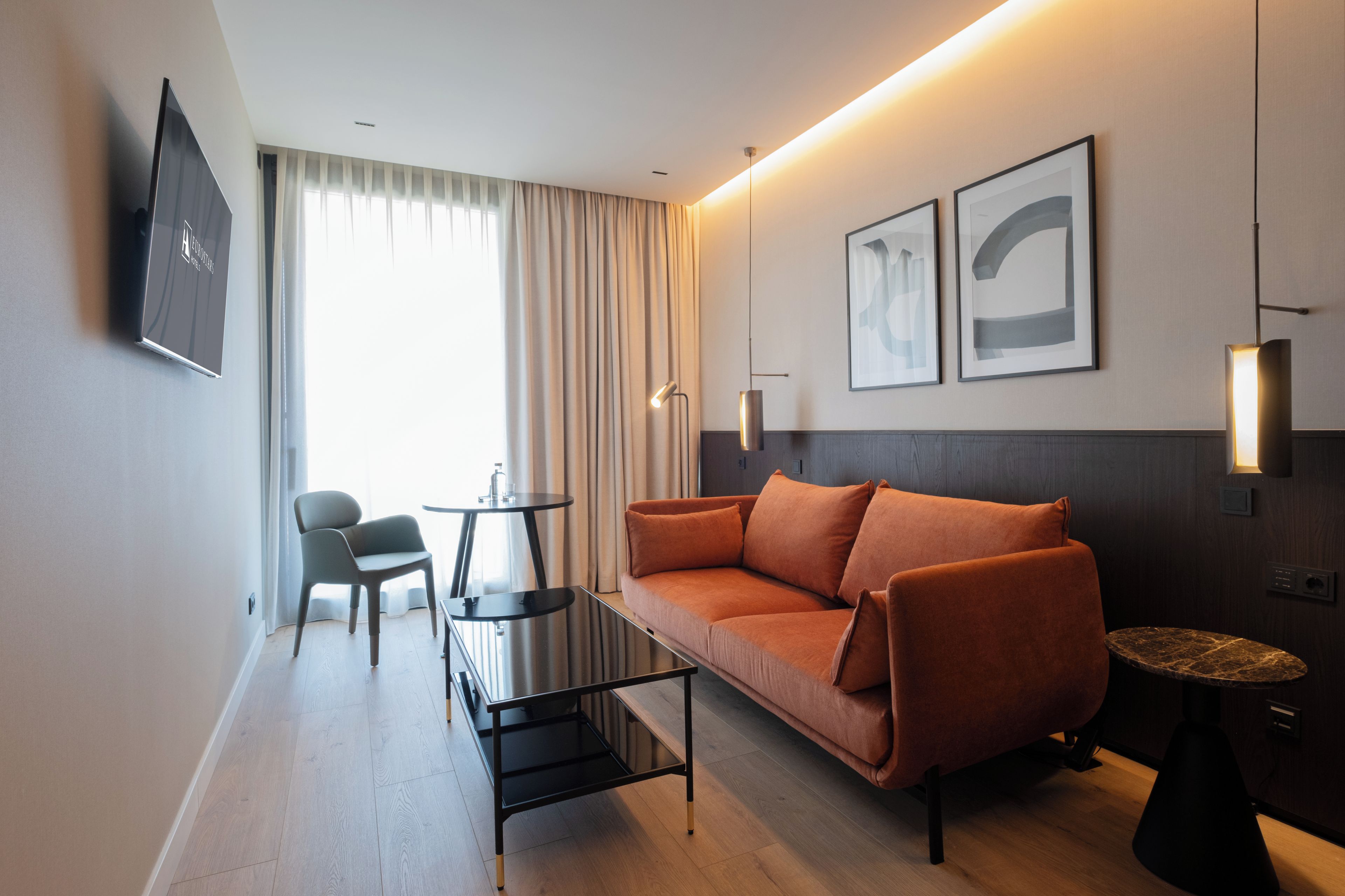 Habitació d'hotel moderna amb un sofà, una taula petita, una cadira i un televisor fixat a la paret.