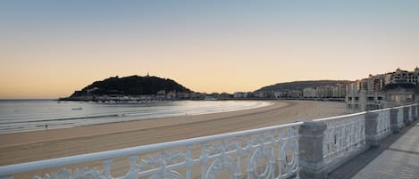 A coastal promenade with a beach, a hill with buildings, and a white ornate railing.