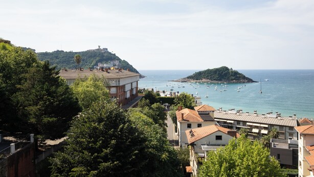 Un pueblo costero con edificios, una construcción en la cima de una colina y una pequeña isla con barcos.