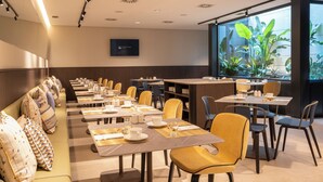 Un comedor de hotel moderno con mesas largas, sillas y un amplio ventanal con plantas.