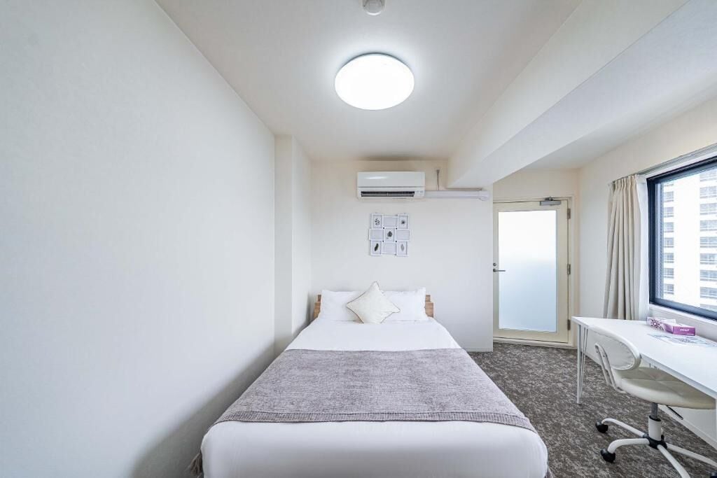 A hotel room with a bed, a desk, an air conditioning unit, and a window.