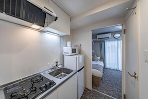 A compact kitchen with a stove, sink, and microwave. A bed with a window and curtains is visible in the adjacent room. - Hoshi noYado Imamiya (Osaka)