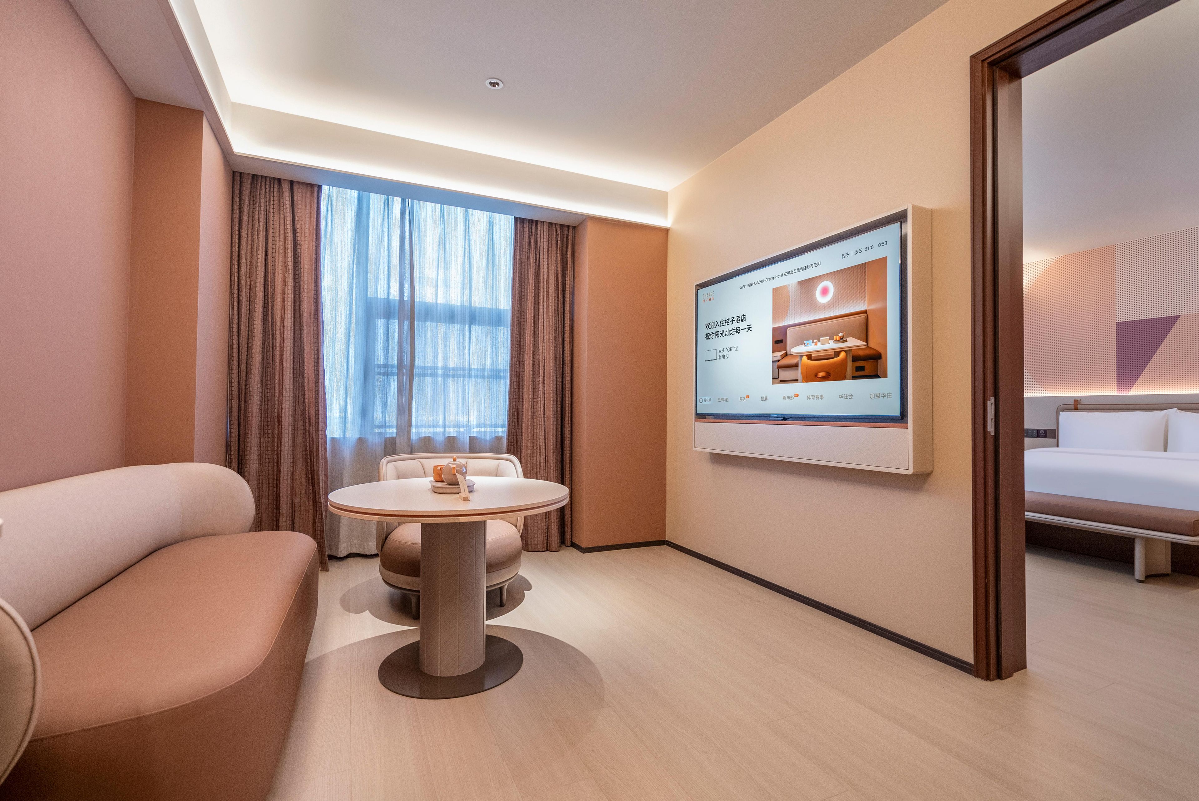 A modern hotel room with a sofa, a round table with a teapot, a bed, and a wall-mounted TV displaying a room image.