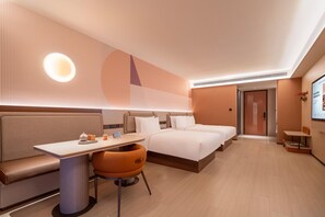 Quarto de hotel moderno com sofá, duas camas de solteiro, uma mesinha com uma cadeira e uma TV grande fixada na parede.