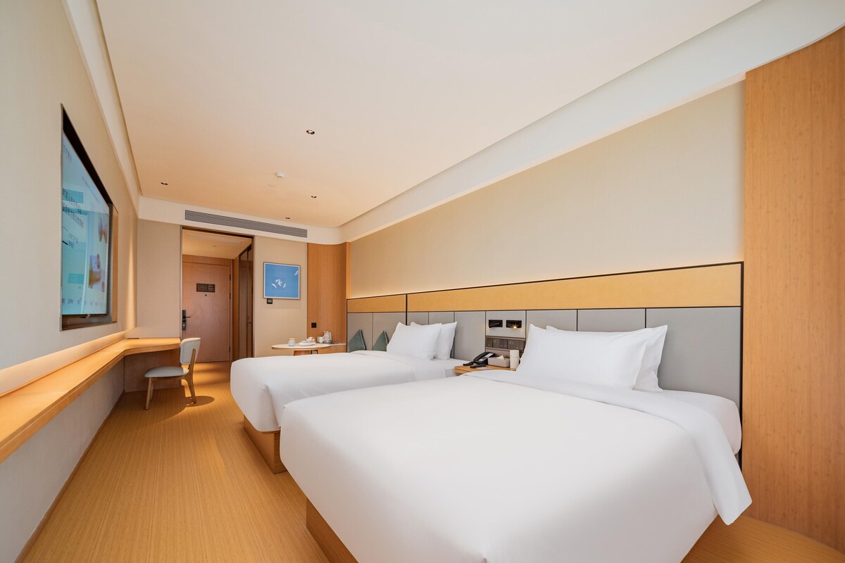A hotel room with two beds, a desk, a chair, and a view of the city.