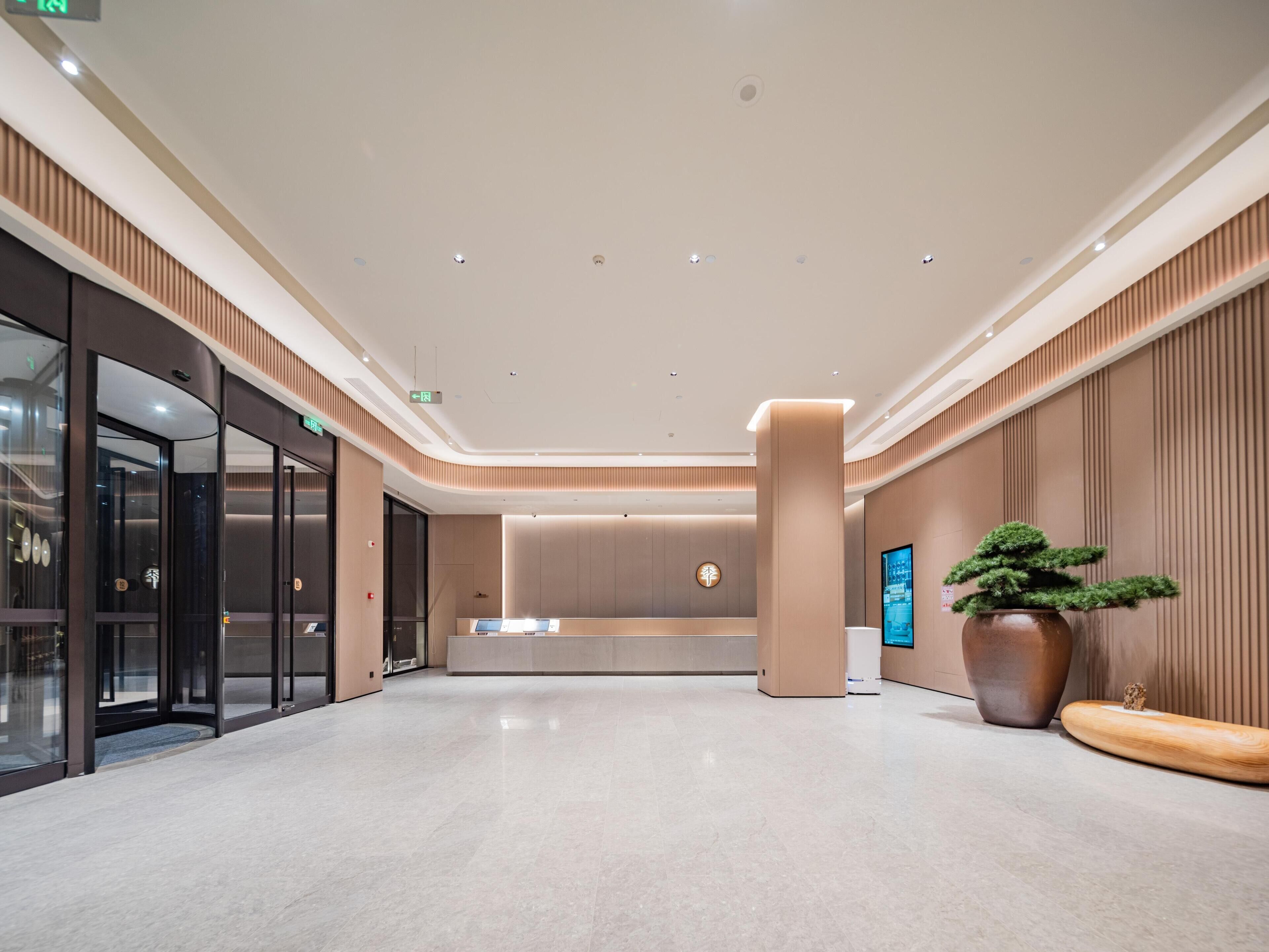 A modern hotel lobby with a reception desk, a digital information screen, and a large potted plant.