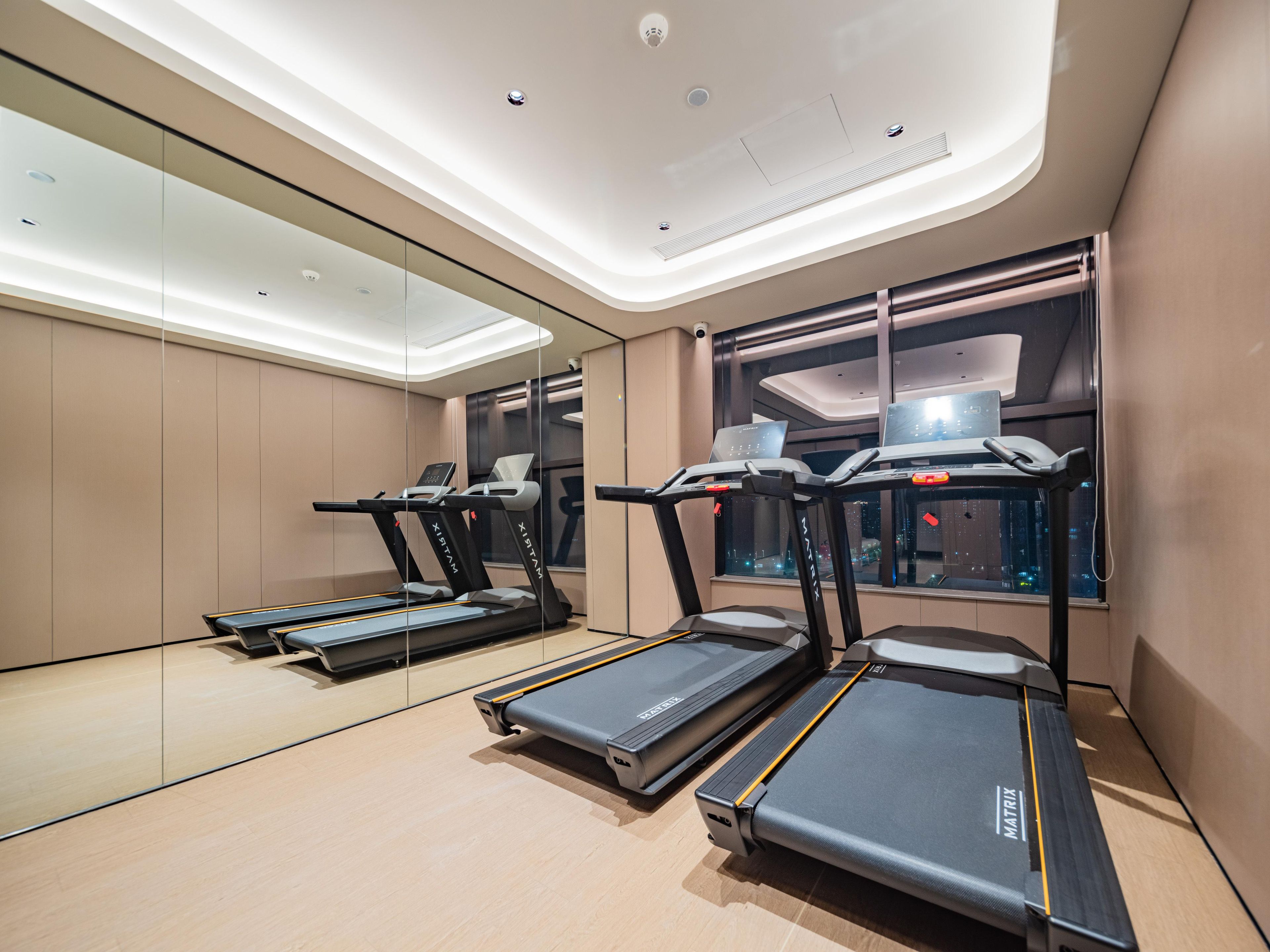 A modern gym with treadmills, a mirrored wall, and a window with a laptop on the desk.