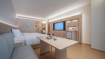 A modern hotel room with a bed, a sofa, a small table with cups, a TV, and a kitchenette.