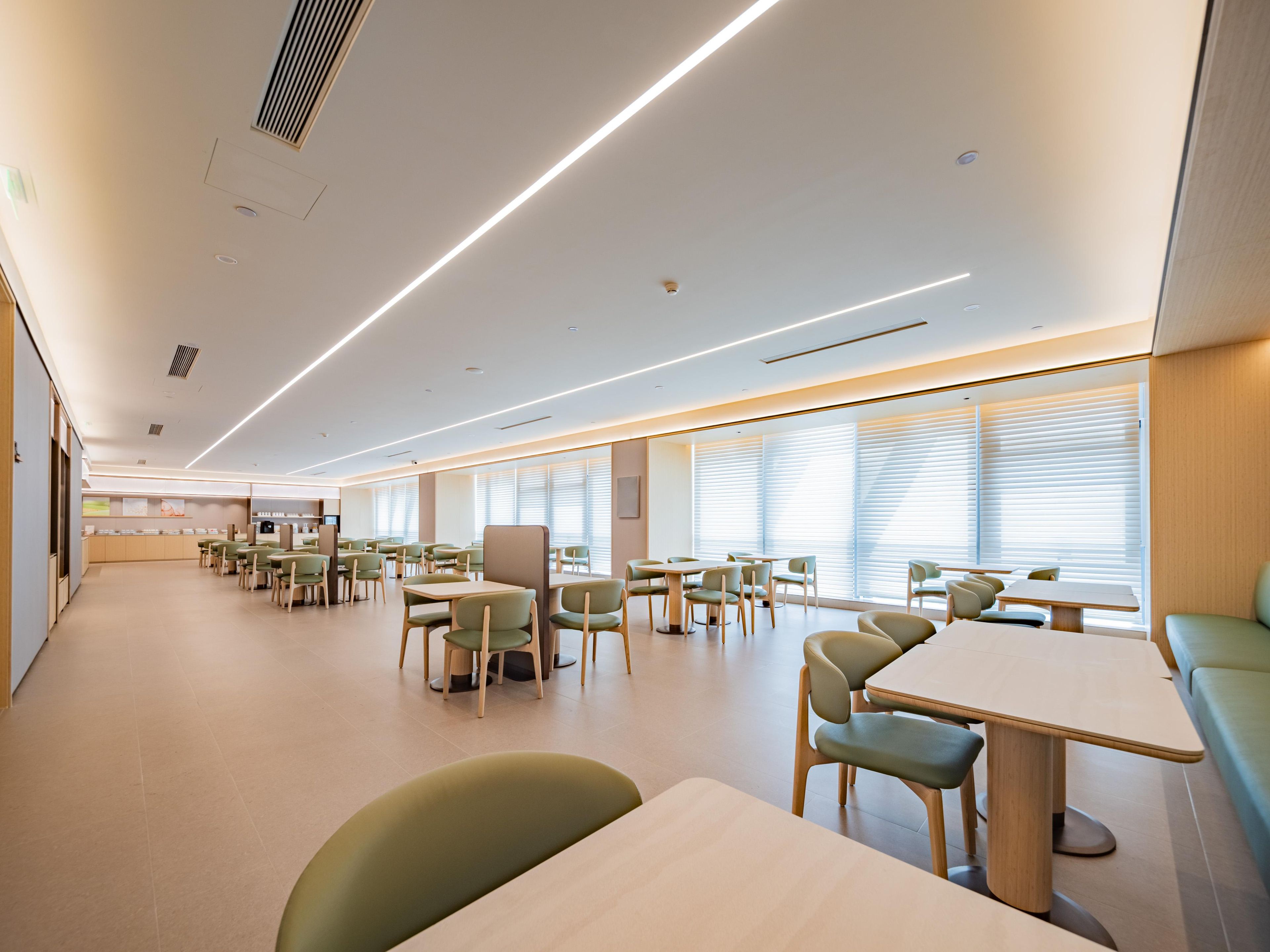 A modern dining area with multiple tables and chairs, a kitchen in the background, and large windows with blinds.
