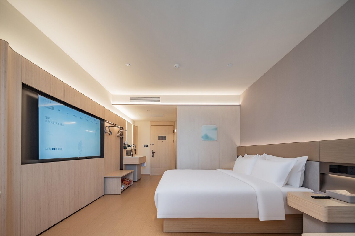A modern hotel room with a large bed, bedside tables, a TV mounted on the wall, and a walk-in closet.