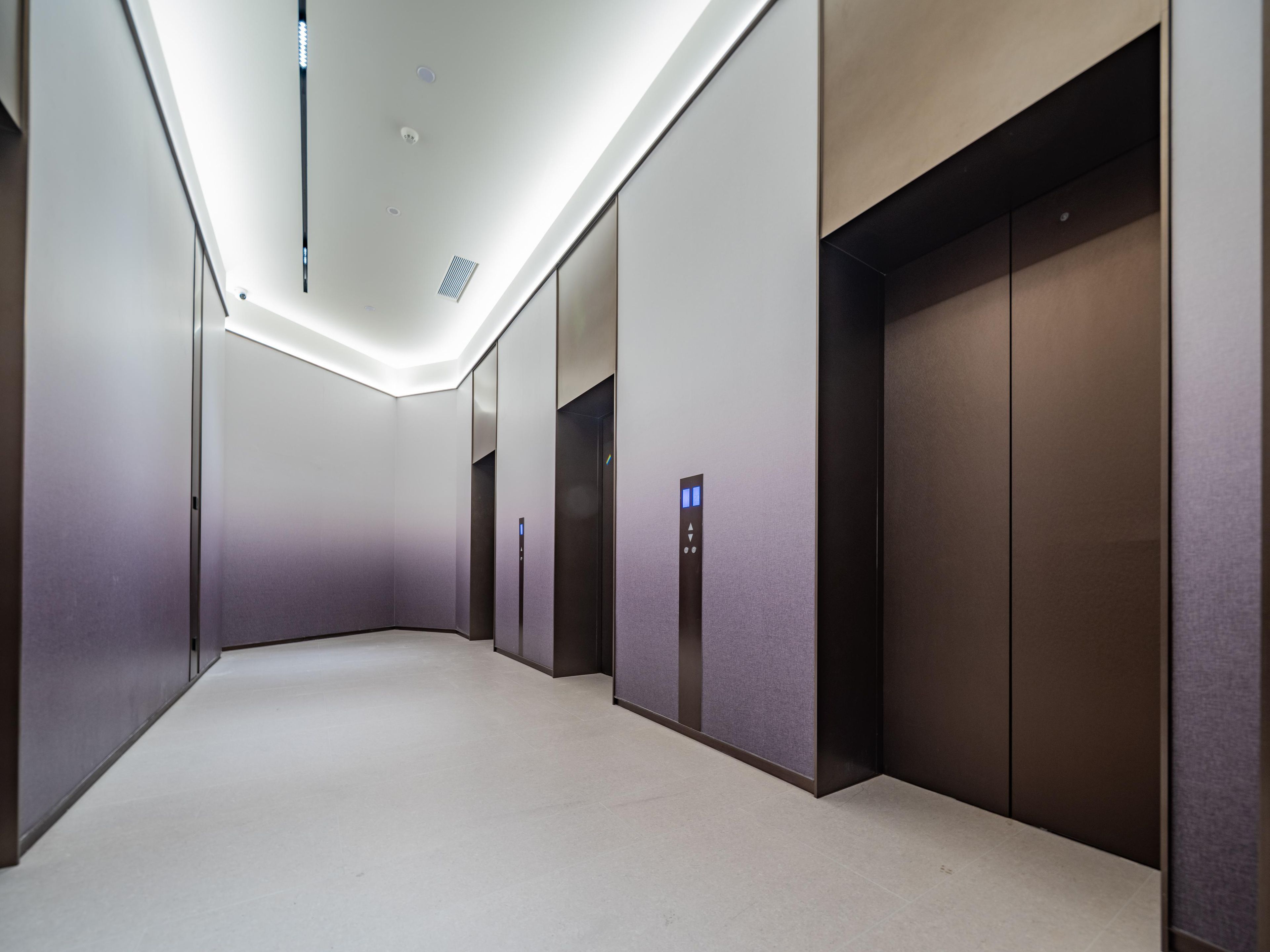 A modern elevator area with a clean, minimalist design, featuring a light-colored floor and walls, and a series of elevators with digital displays.