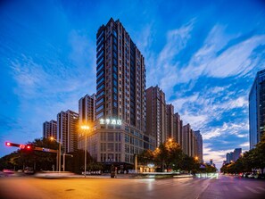 Un edificio de gran altura con varios pisos, una entrada destacada y un cielo despejado con nubes.