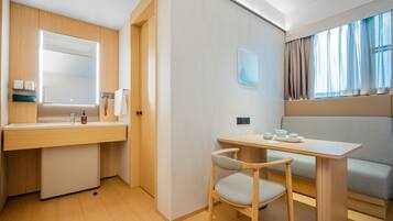 A modern hotel room with a wooden vanity, a small table with a chair, a sofa, and a window with curtains.