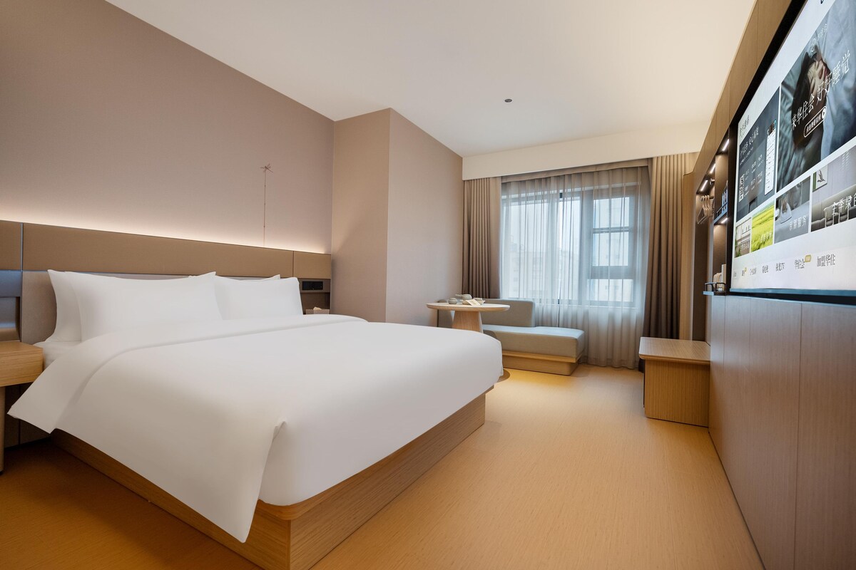 Habitación de hotel con una cama grande, una mesita, un sofá y una pantalla de televisión mostrando un menú.