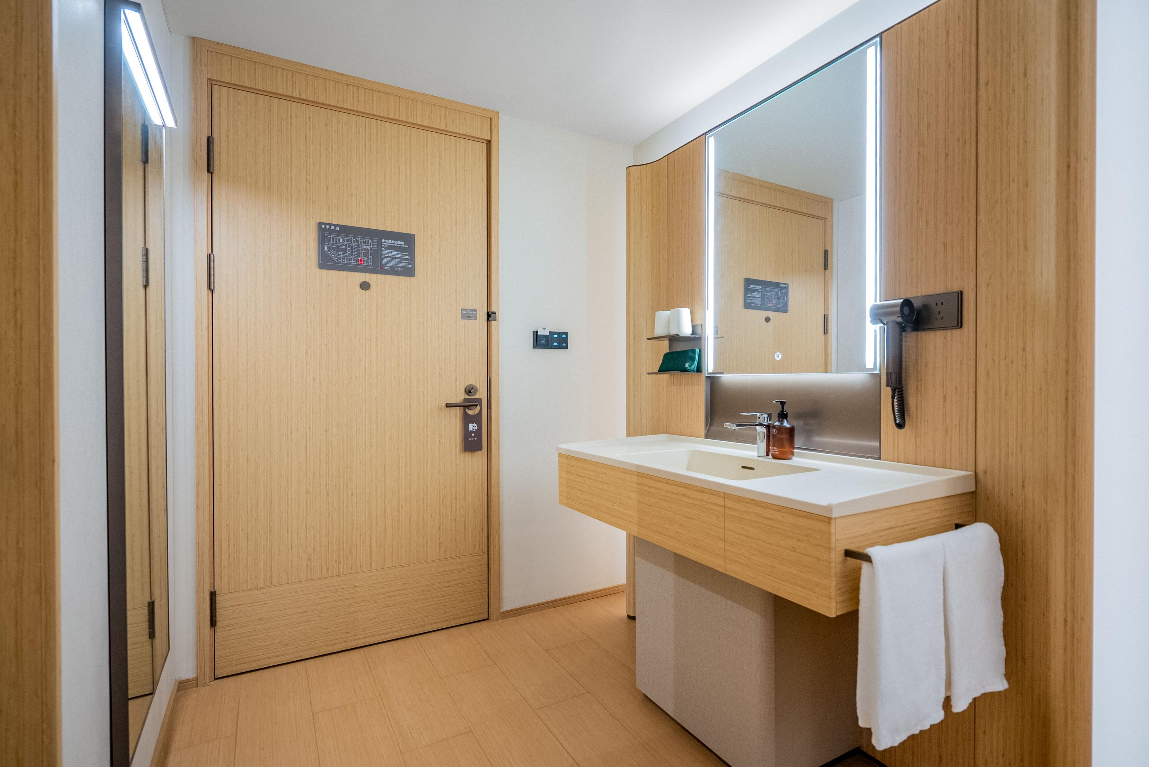 A hotel bathroom with a wooden door, a large mirror, a sink with a towel, and a toilet.